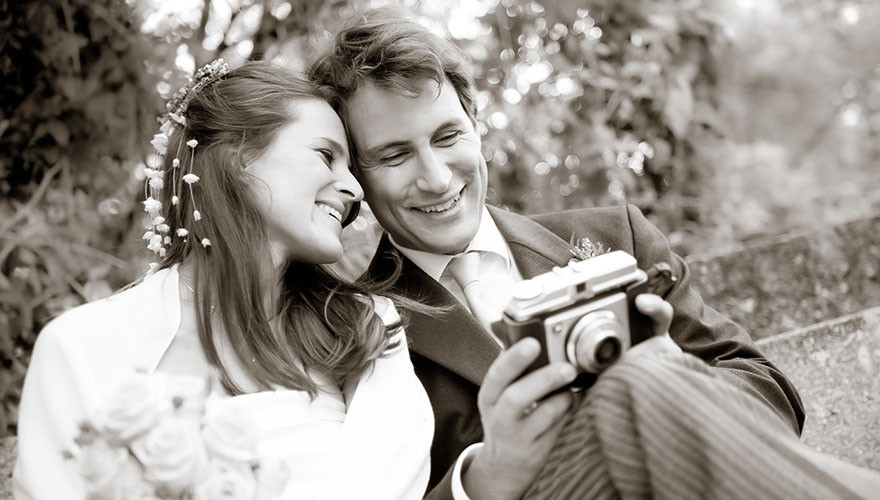 Ein Bräutigam zeigt seiner Braut Bilder auf seiner Kamera. Die Braut presst ihre Stirn an seine Schlefe und lacht. Der Bräutigam lacht ebenfalls.
Sie sitzen auf einer Bank in einem Park. Im Hintergrund erkennt man Bäume.