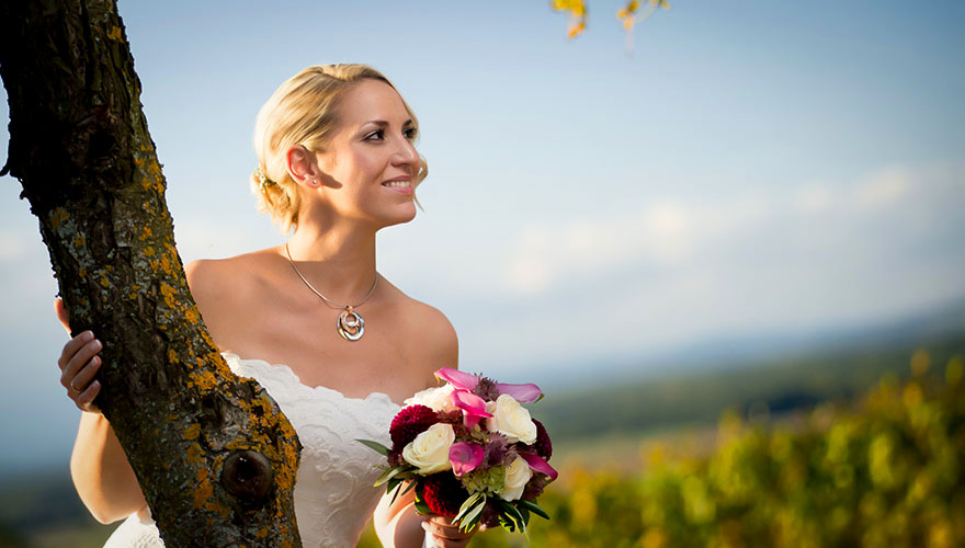 Eine Braut steht bei einem Baum und blickt in den herbstlichen Weingarten.