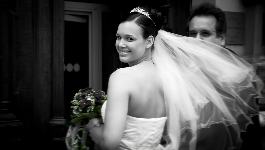 Eine Braut durchschreitet mit ihrem Vater den Eingang zum Standesamt. Der Wind bläst den Schleier vor das Gesicht des Vaters.