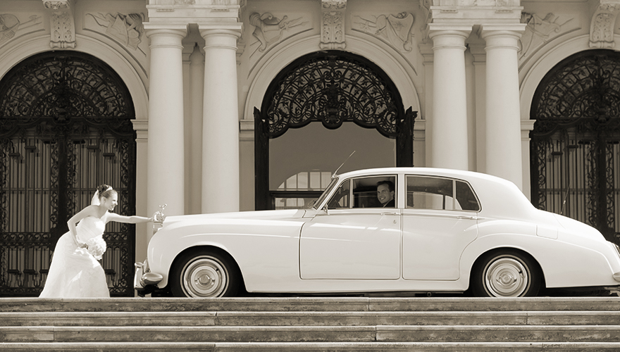 Vor dem Schloss Belvedere steht ein alter Rolls Royce in dem ein Bräutigam hinter dem Lenkrad sitzt. Draussen steht die Braut und stemmt sich gegen den Wagen, sodass er nicht weiterfahren kann.