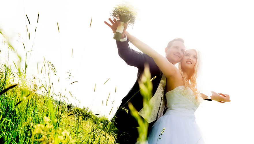 Auf einer sommerlichen Wiese mit hoch gewachsenen Grashalmen steht ein Bräutigam hinter seiner Braut. Er hält Ihre Arme weit ausgestreckt. Es hat den Anschein als würden sie fliegen.