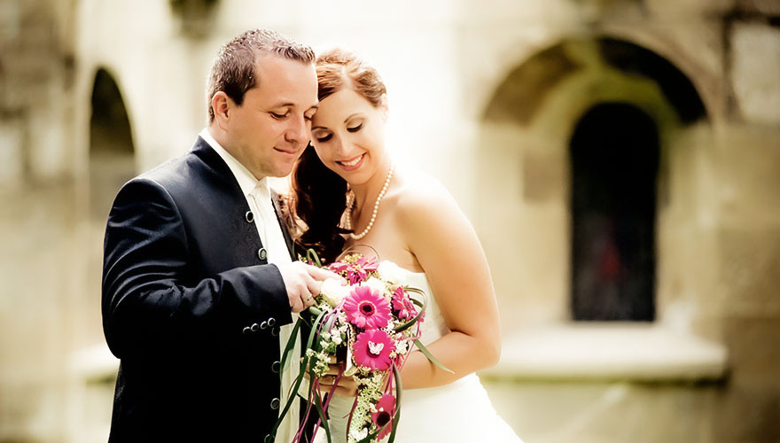 Ein Bräutigam zeigt seiner Braut den Brautstrauss. Ihre Stirn berührt seine Wange, sie lächelt dabei. Im Hintergrund erahnt man die Kirche.