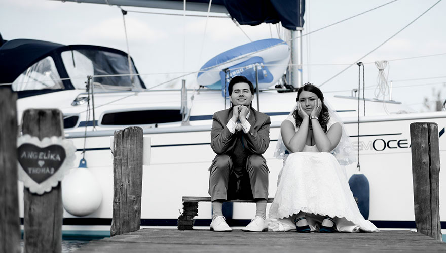 Auf einem Steg am Neusiedlersee sitzt ein Brautpaar und hat die Köpfe in die Hände gelegt. Sie beobachten den Fotografen. Im Hintergrund fährt ein Segelschiff sehr dicht am Steg vorbei.