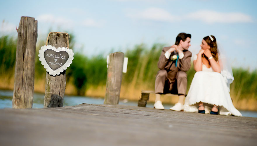 Ein Brautpaar sitzt am Steg vom Neusiedlersee und konzentriert sich nur auf sich selber. Im Vordergrund ist auf einem Pfosten ein Schild angebracht, dort stehen die Namen Angelica und Thomas.