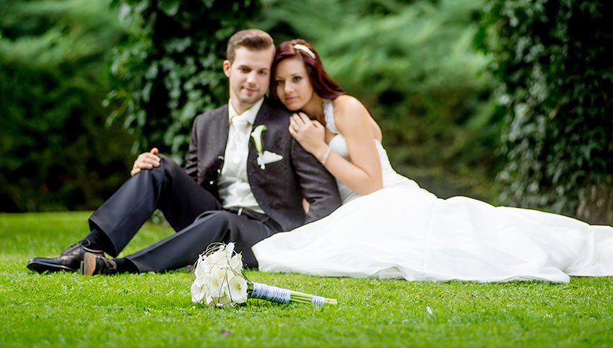 Ein Brautpaar sitzt auf einer saftigen Wiese. Etwas weiter vor ihnen liegt Ihr Brautstrauss. Beide betrachten den Blumenstrauss.