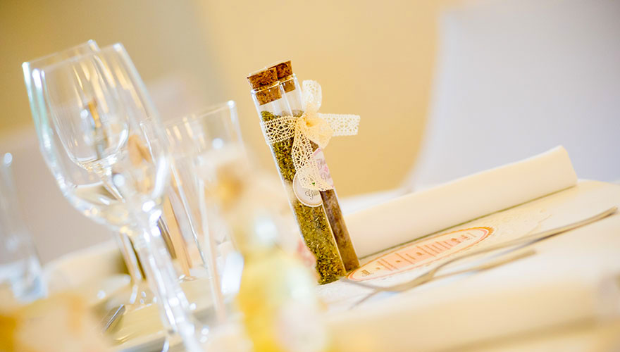 Ein detailierter Blick auf einen Tisch einer Hochzeitstafel. Neben Gläsern und Servieten gib es als Gastgeschenk Gläser mit Gewürzen.