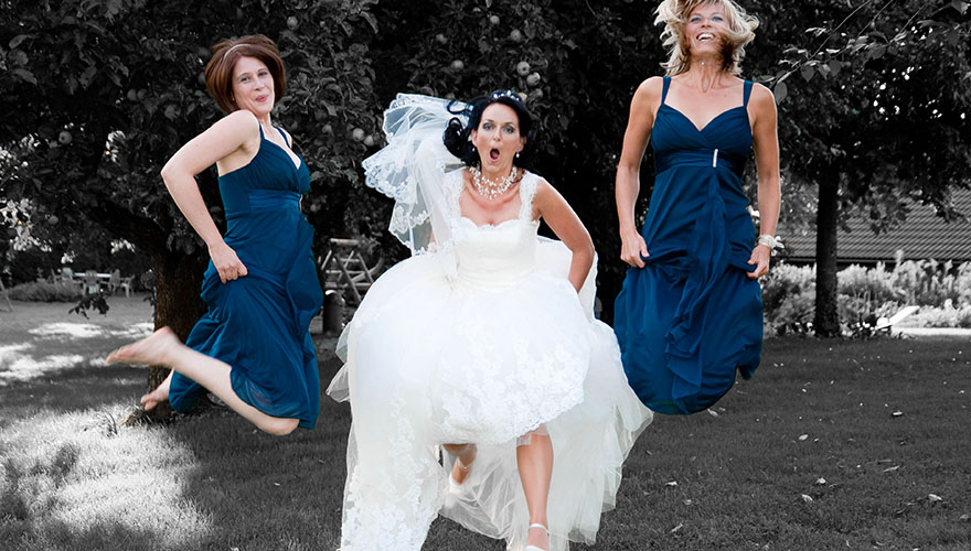 In einem Garten springen eine Braut und ihre zwei Brautjungfern sehr hoch in die Luft. Die Brautjungfern lachen und die Braut hebt Ihr Kleid beim Sprung um nicht über Ihre Schleppe zu stolpern.