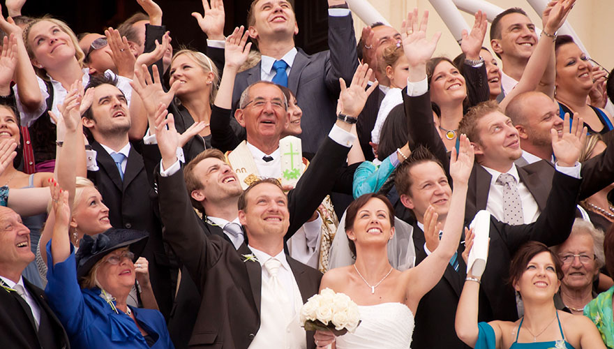 Eine Hochzeitsgesellschaft, mitten drinnen das Brautpaar. Etwa 25 Hochzeitsgäste blicken in den Himmel, strecken die Hände in die Höhe und lachen laut.