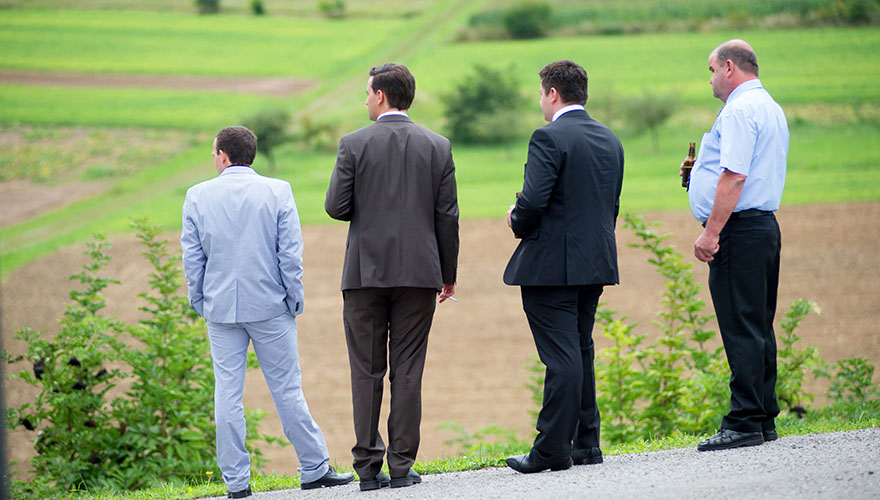 Vier Männer auf einer Hochzeit blicken von einem Hügel ins Tal. Ein Gast raucht eine Zigarett und ein anderer trinkt ein Bier.