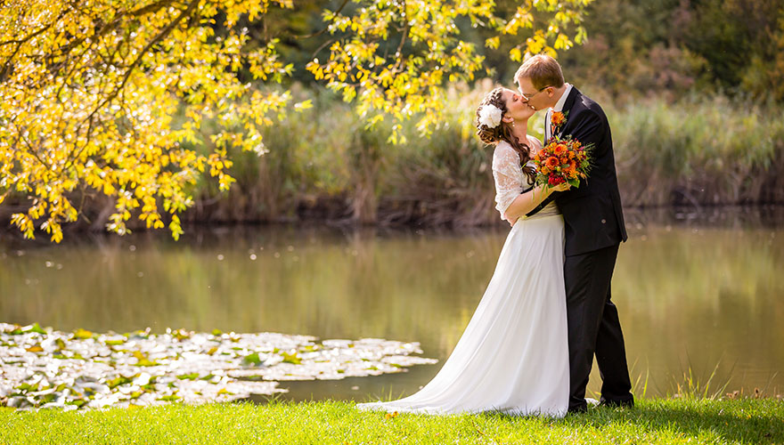 An einem Teich steht ein Brautpaar. Der Bräutigam küsst seine Braut.