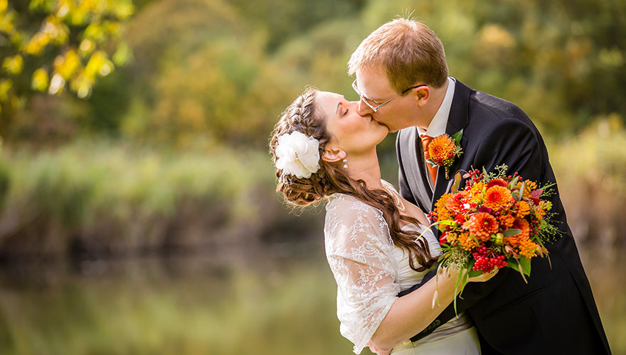 Ein Bräutigam küsst seine Braut. Im Hintergrund erkennt man einen Teich. Es ist Herbst.