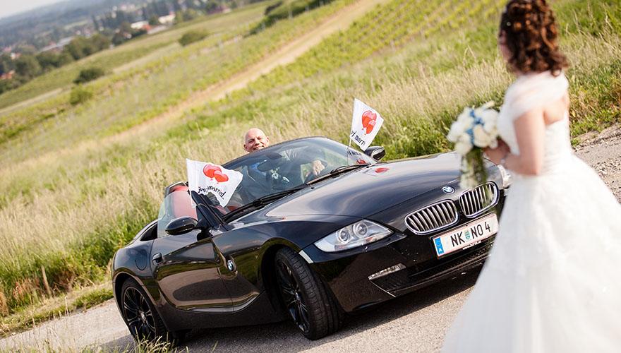 Auf einer Landstrasse fährt ein Bräutigam in einem Caprio. Das Caprio ist mit zwei Fahnen geschmückt auf denen Herzen aufgezeichnet sind. Die Braut erwartet das Auto.