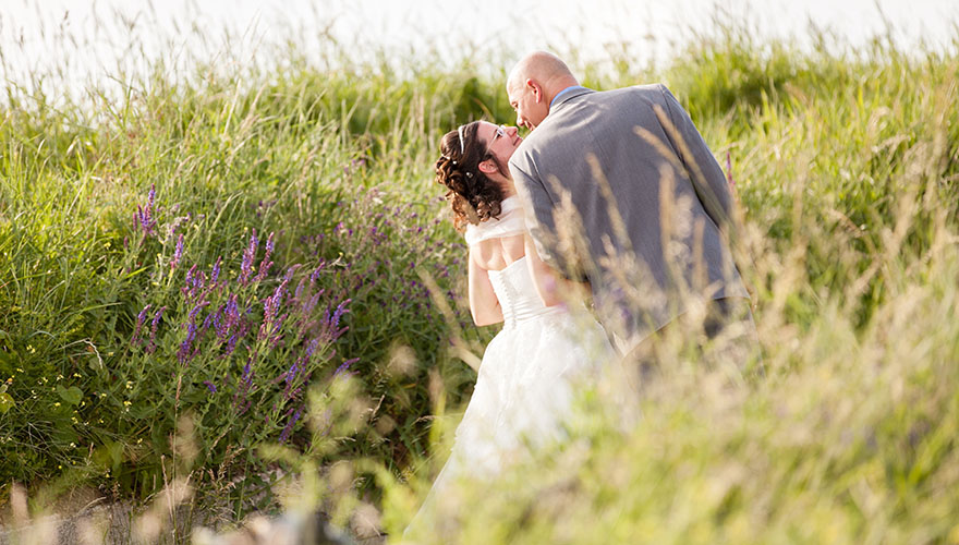 Auf einem Feldweg, mitten in sehr hohem Gras spaziert ein Brautpaar. Es scheint, als würden sie sich gleich küssen.