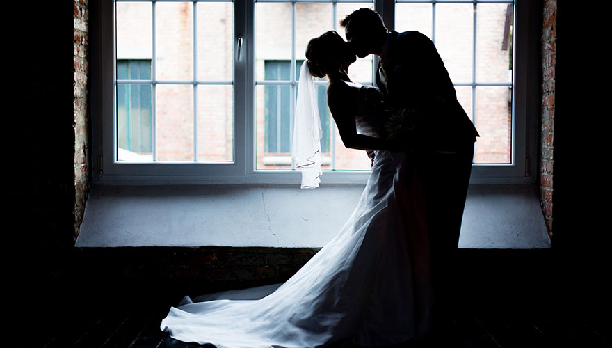 Vor einem grossen Fenster hält ein Bräutigam seine Braut ganz fest. Sie beugt sich etwas nach hinten und er küsst sie von oben auf den Mund.