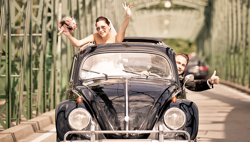 Ein Brautpaar fährt in einem alten Käfer, ein Volkswagen aus den 60er Jahren. Die Braut streckt Ihren Kopf durch das offenene Autodach. Der Bräutigam fährt den Wagen und streckt seinen Kopf durch die Seitenscheibe und zeigt mit dem Daumen nach oben. Si