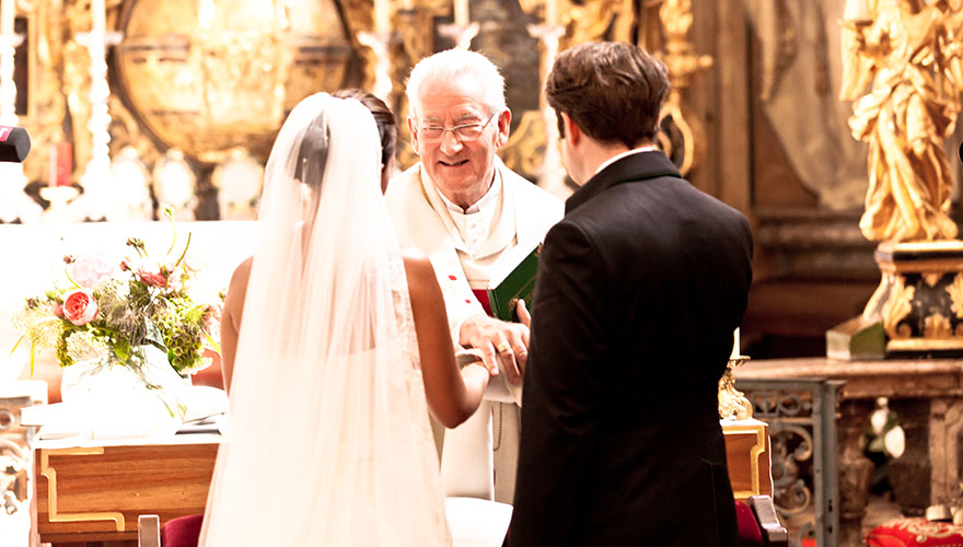 In einer Kirche steht ein Brautpaar vor dem Priester. Das Brautpaar hält sich an den Händen, der Priester greift nach den beiden Händen und lächelt die Beiden an.