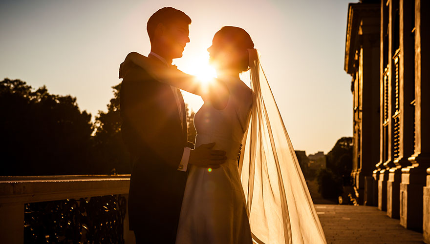 Es ist kurz vor Sonnenuntergang auf einer Terasse eines Schlosses oder Palais. Eine Braut legt Ihre Hände auf die Schultern des Bräutigams und sieht in seine Augen. Er fasst die Braut an Ihrer Hüfte und lächelt sie an.