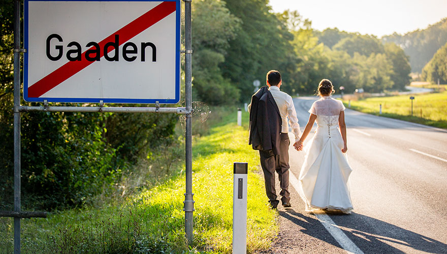 Ein Brautpaar geht eine Strasse entlang. Am Strassenrand steht eine Verkehrstafel auf der das Ende des Ortes: Gaaden angezeigt wird. Die kurvenreiche Strasse verläuft im Hintergrund im Wald. Das Brautpaar hält sich an den Händen, beide sehen nach vorne.