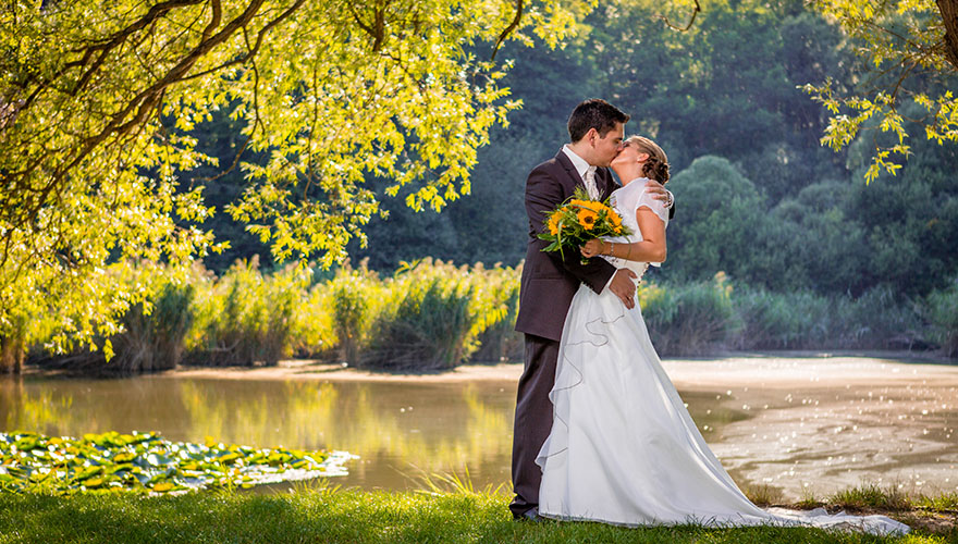 Eine herstliche Landschaft. Vor einem Teich hält ein Bräutigam seine Braut und zieht sie an sich. Er küsst sie leidenschaftlich.