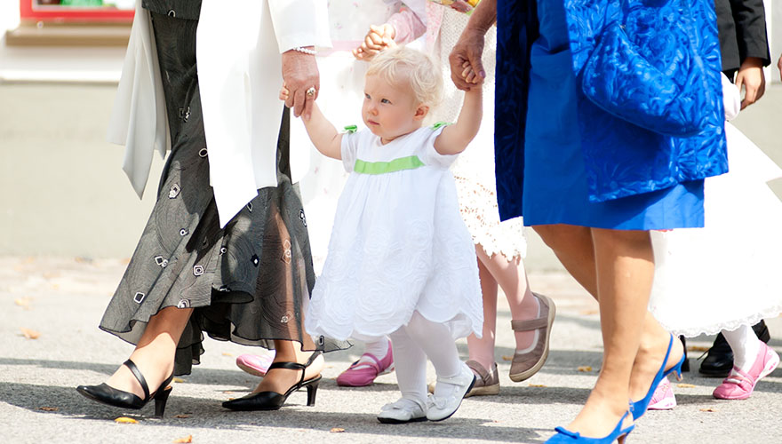 Ein kleines Mädchen mit ca. 1 Jahr geht zwischen zwei Frauen die es an den Händen halten. Sie gehen zu einer Hochzeit und alle sind sehr festlich gekleidet.