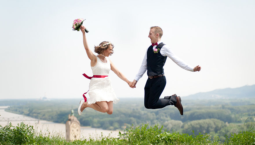 Ein frisch vermähltes Hochzeitspaar mit jugendlichem Outfit, die Braut trägt ein knielanges Kleid und der Bräutigam eine Anzugweste ohne Sakko, springen hoch in die Luft. Hinter ihnen liegt die Wachau durch die die Donau fliest.