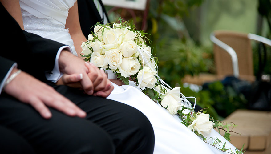 Ein Brautpaar sitzt am Standesamt und hält sich ganz fest an den Händen. Die Braut hat in ihrer zweiten Hand einen Blumenstrauss mit vielen Rosen.