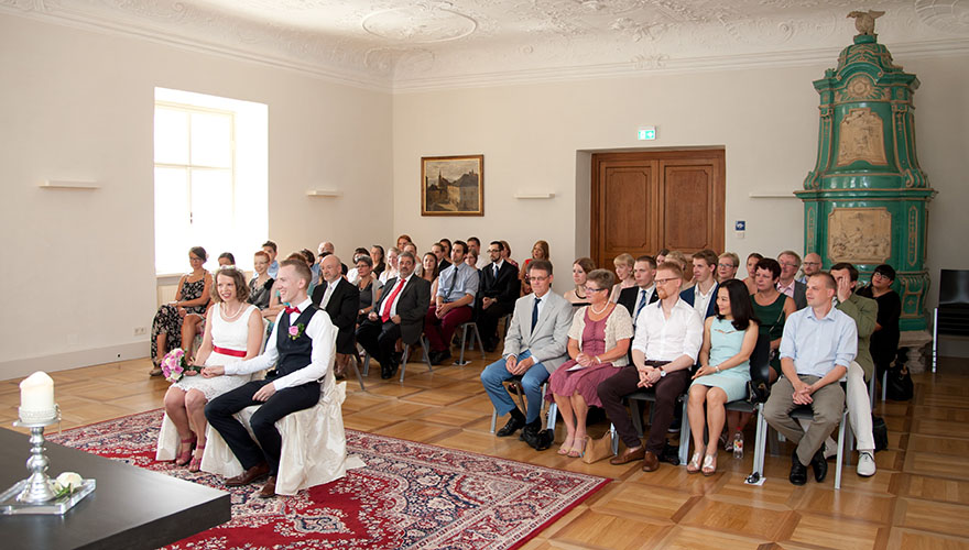 In einem Rathaussaal sitzt eine ganze Hochzeitsgesellschaft. Das Brautpaar ist ganz konzentriert auf die Trauung und lächelt verschmilzt.