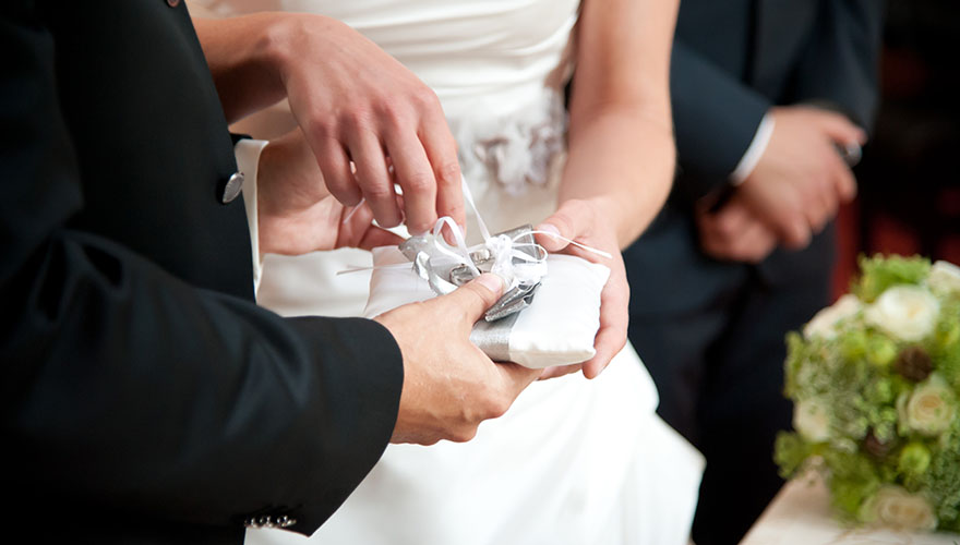 Ein Bräutigam hält ein Ringkissen und die Braut öffnet die Bänder an denen die weissgoldenen Ringe befestigt sind. In Kürze wird sie ihrem Bräutigam den Ring anstecken.