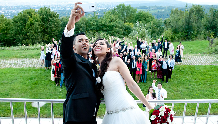 Ein Brautpaar steht auf einer Terasse. Im Hintergrund auf der Wiese steht die ganze Hochzeitsgesellschaft. Das Brautpaar macht mit Ihrem Handy ein Selfie und grinsen um die Wette.