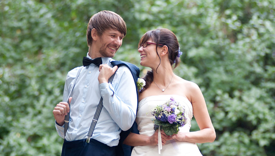 Ein Brautpaar steht vor einem Gebüsch. Der Bräutigam hat sein Sakko ausgezogen. Es hängt lässig über seine Schulter. Er dreht den Kopf zu seiner Brau. Sie lächelt ihn an während sie mit beiden Händen ihren Brautstrauss festhält.