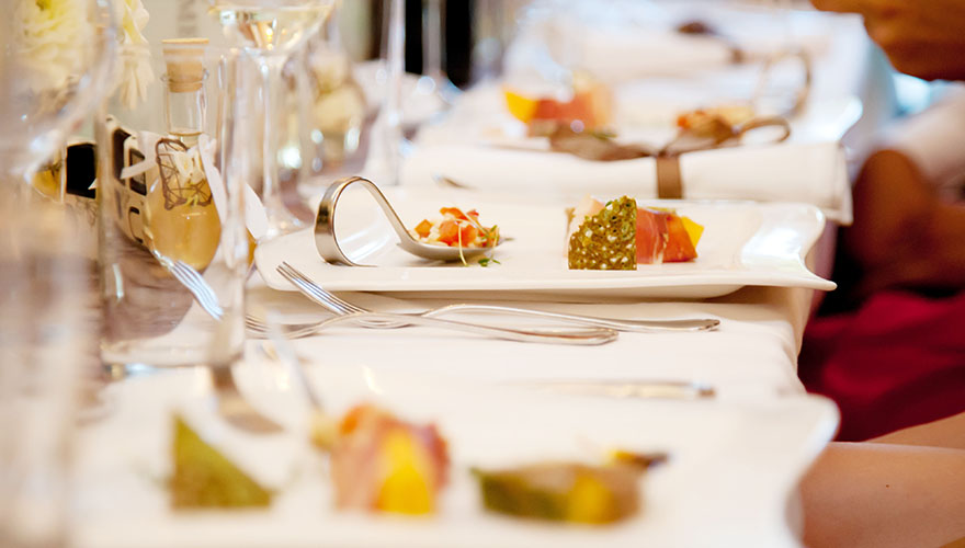 Bei einer Hochzeit wurde die Vorspeise serviert. Auf eckigen Tellern stehen gebogene Löfel mit Tomaten. Rohschinken und gitterförmiges Backwerk stehen daneben.