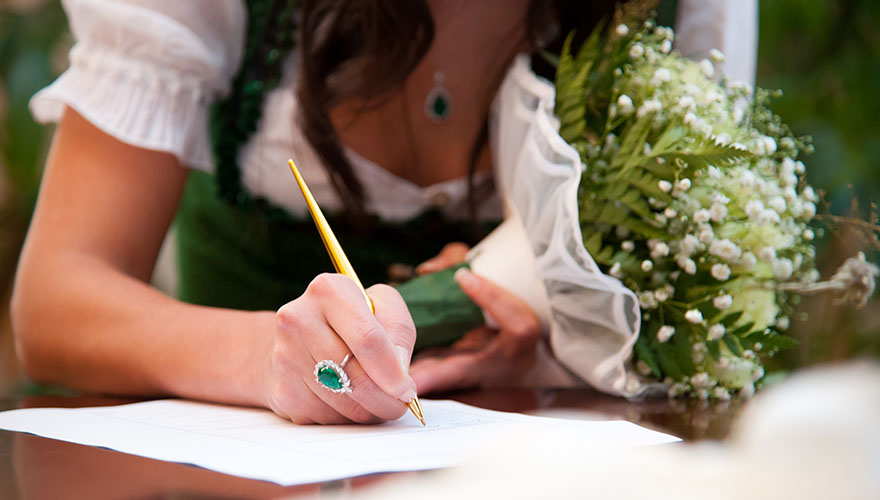 Eine Brau unterschreibt im Ehebuch beim Standesamt. Sie trägt ein Dirndl und hält in der zweiten Hand ihren Brautstrauss. Auf der Hand mit der sie unterschreibt ist auch der Verlobungsring mit einem grossen Edelstein zu erkennen.