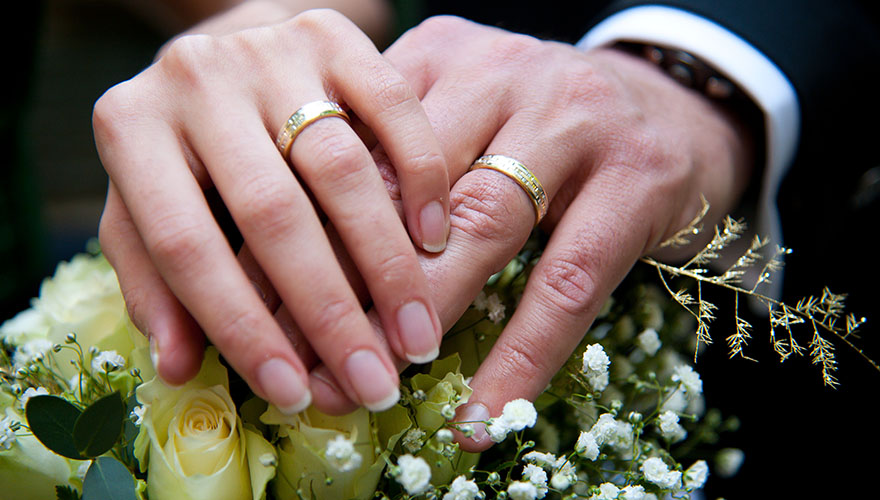 Ein Brautpaar präsentiert seine reichlich verzierten, goldenen Eheringe. Dafür halten sie die Hände auf den Brautstrauss und die Braut legt ihre Hand über die Hand des Bräutigams.