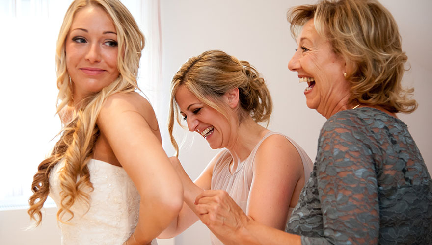 Zwei Frauen helfen einer Braut in ihr Brautkleid. Sei lachen dabei viel. Die Braut presst Ihre Corsage zusammen.