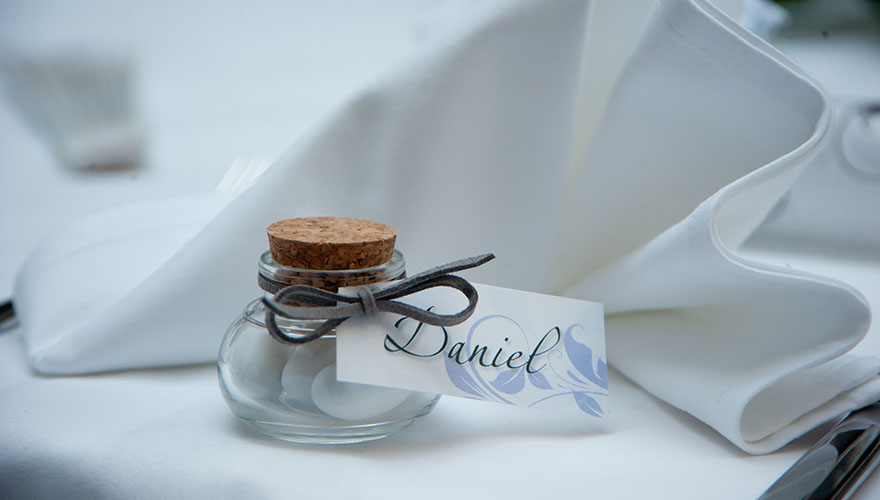 Bei einer Hochzeit steht ein Glas gefüllt mit Bonbons vor einer gefalteten Serviette. Am Glas hängt ein Schild mit einer Tischnamenskarte.