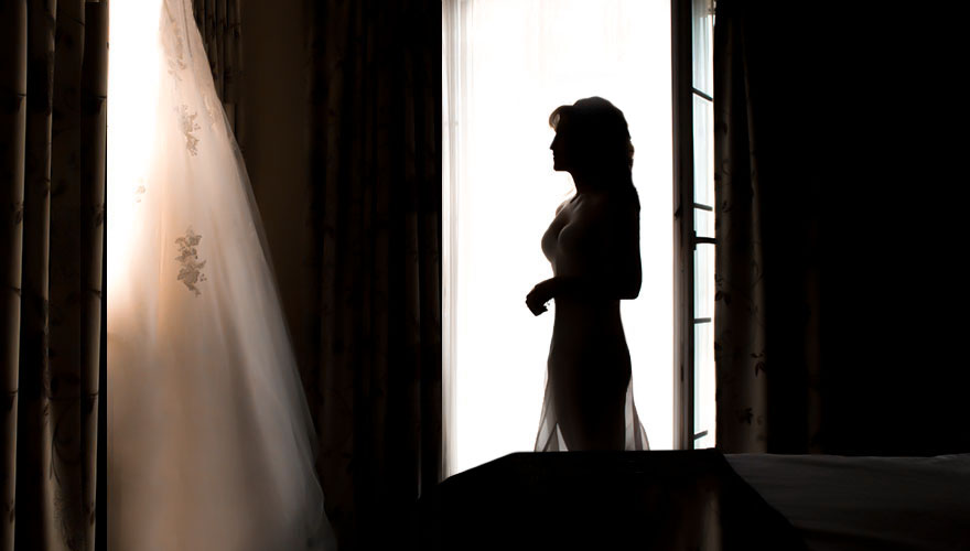 In einem dunklen Raum, nur die Fenster zeigen das es Tag ist, steht eine Braut in Unterwäsche und Unterkleid. Sie betrachtet Ihr Brautkleid welches am Fenster hängt.