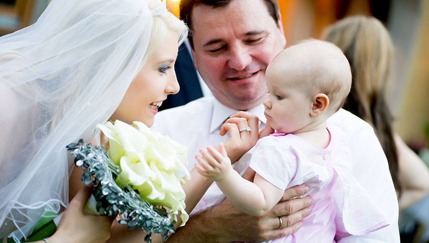 Bei einer Hochzeit präsentiert ein stolzer Vater seine kleine Tochter, welche er im Arm hält, der Braut. Sie streichelt dem Kind das Kinn.