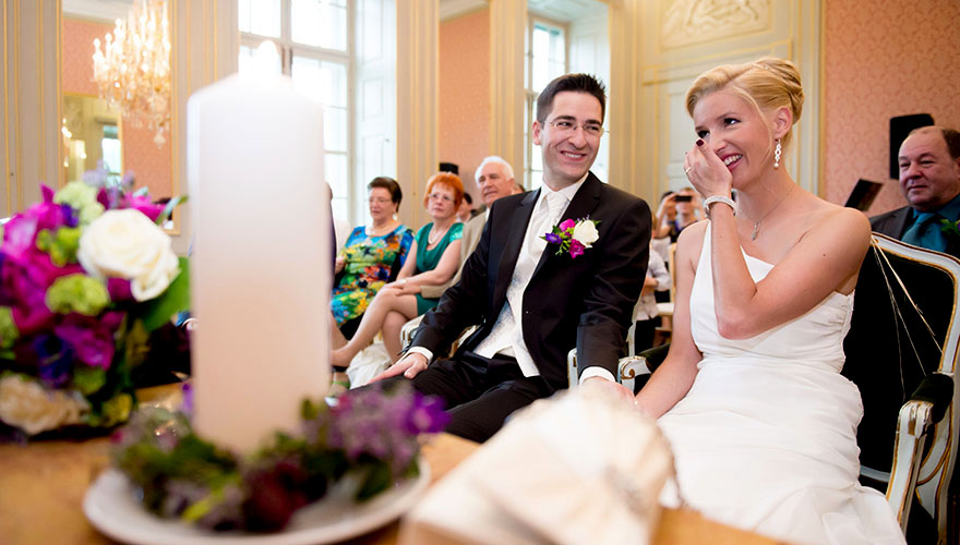 Hinter der Hochzeitsgäste bemerkt man das Brautpaar vor seinen Hochzeitsgästen. Weil die Braut Tränen in den Augen hat, lächelt sie der Bräutigam liebevoll an.
