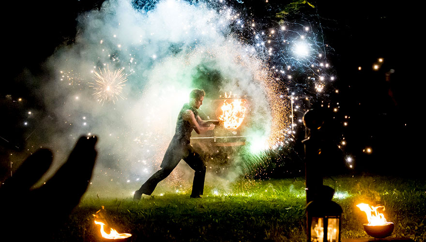 Es ist Nacht bei einer Hochzeitsfeier. Ein Artist schwingt Ketten mit Feuerwerkskörpern. Überall sprühen Funken. Im Vordergrund sieht man Tellergrosse Feuerschalen mit lodernden Flammen.