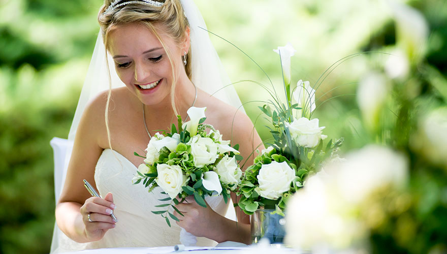 Eine Braut sitzt in einem Garten am Tisch und setzt ihre Unterschrift ins Ehebuch. Sie lächelt und hält ihren Blumenstrauss ganz fest.