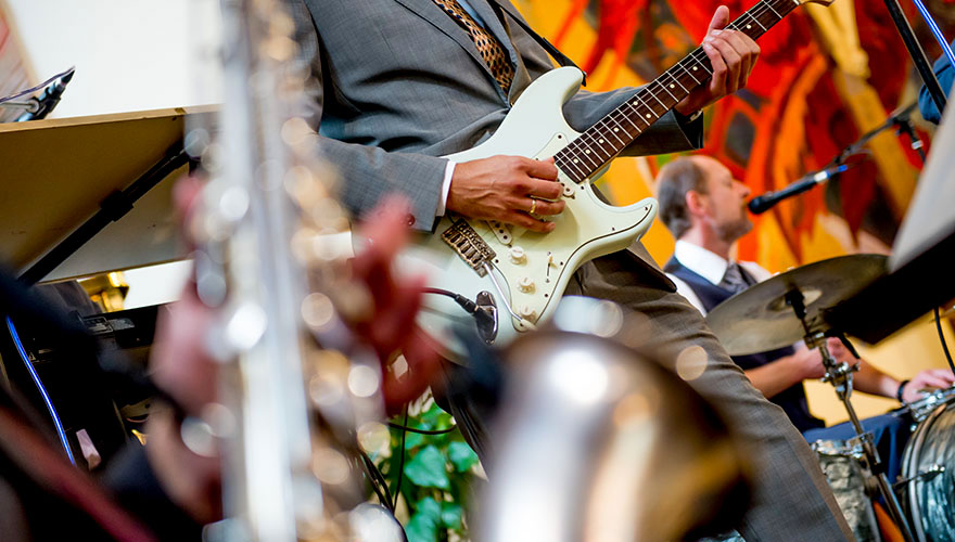 Zu einer Hochzeit spielt in der Kirche eine Band auf. Der Gitarist zupft Saiten und im Vordergrund sieht man wie ein Saxophon gespielt wird. Im Hintergrund singt der Schlagzeuger ins Mikrofon.