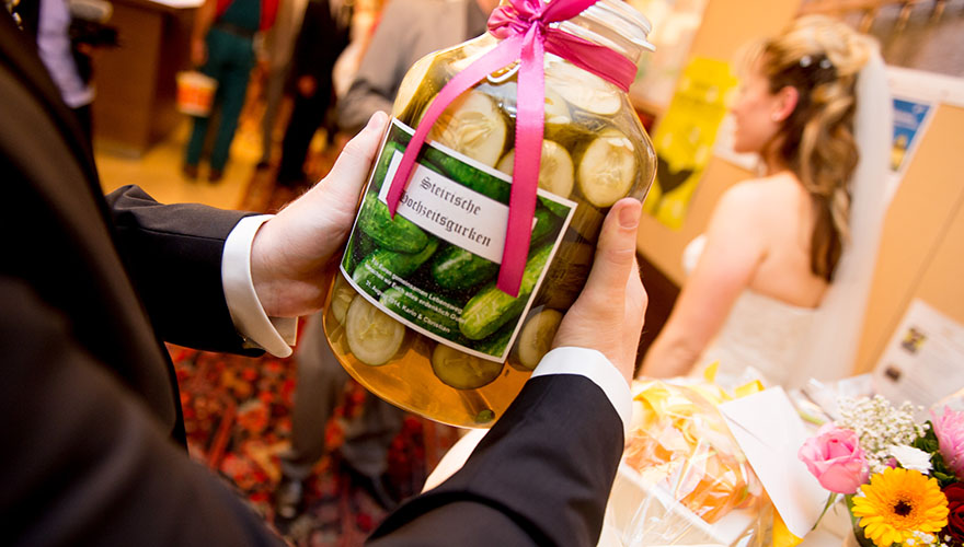 Während Gäste einer Braut zu ihrer Hochzeit gratulieren und Geschenke überreichen, hält ein Mann ein Geschenk in der Hand. Es handelt sich um ein grosses Grukenglas voll mit Gruken in Essig oder Salz. Auf dem Etikett steht: Steirische Hochzeitsgruken.