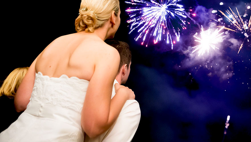 Am nächtlichen Himmel explodieren Feuerwerkskörper. Ein Brautpaar steht und betrachtet das Spektakel am Himmel.