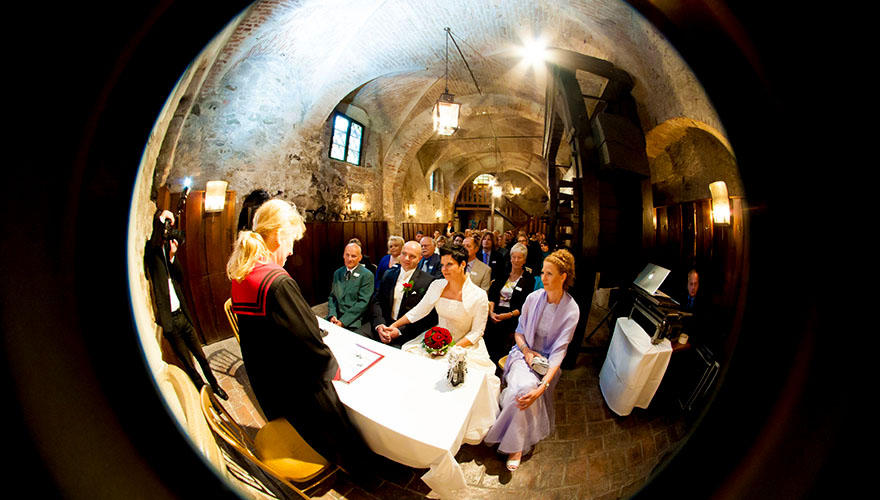 In einem engen Keller findet eine Hochzeit statt. Der Fotograf hat schwierigkeiten genug Platz zu finden um alle Gäste und das Brautpaar auf ein Foto zu bekommen.