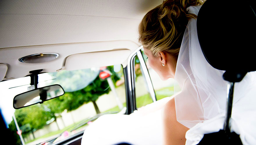 Eine Braut sitzt am Beifahrersitz im Auto und blickt auf die Strasse. Das Auto fährt durch eine Allee.