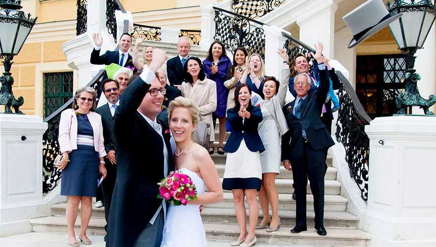 Eine Hochzeitsgruppe steht auf den Vordertreppen von Schloss Schönbrunn und klatscht dem Brautpaar im Vordergrund zu. Der Bräutigam schmeisst seinen Zylinderhu in die Luft.