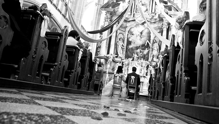 In einer Kirche findet eine Hochzeit statt. Man erkannt den Steinboden auf dem noch Blüttenblätter vom Einziehen liegen. Das Brautpaar sitzt am Ende des Gangs und hält sich an den Händen.